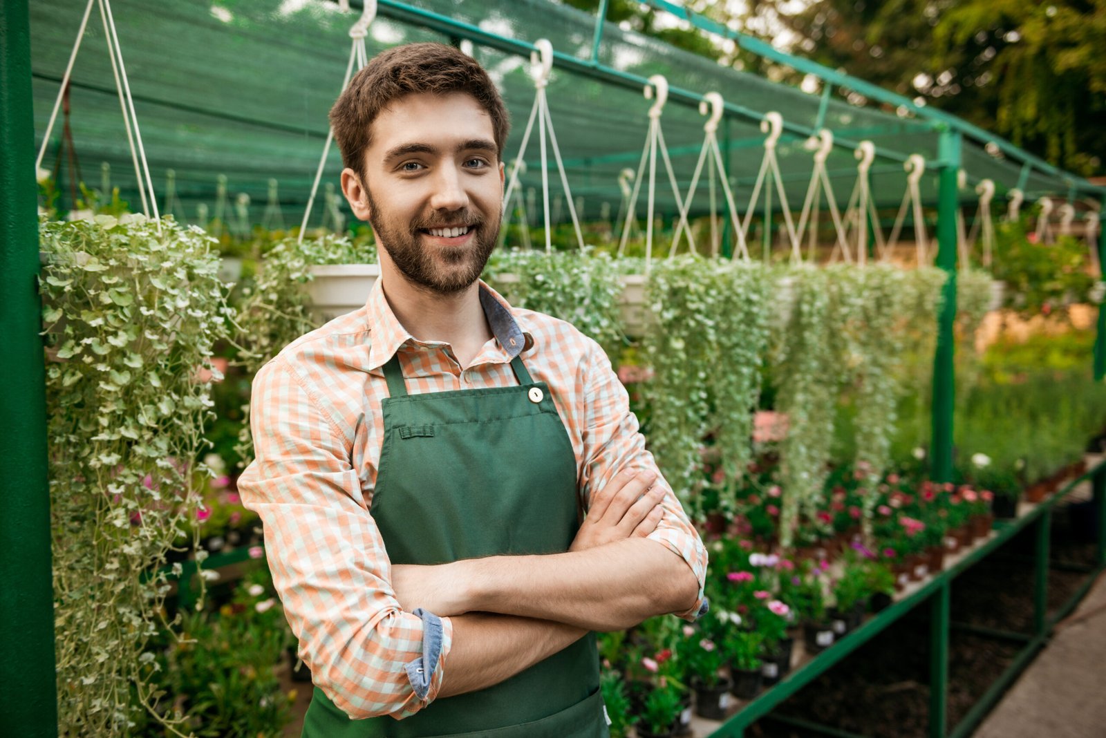 Young handsome gardener smiling, posing with crossed arms among flowers.